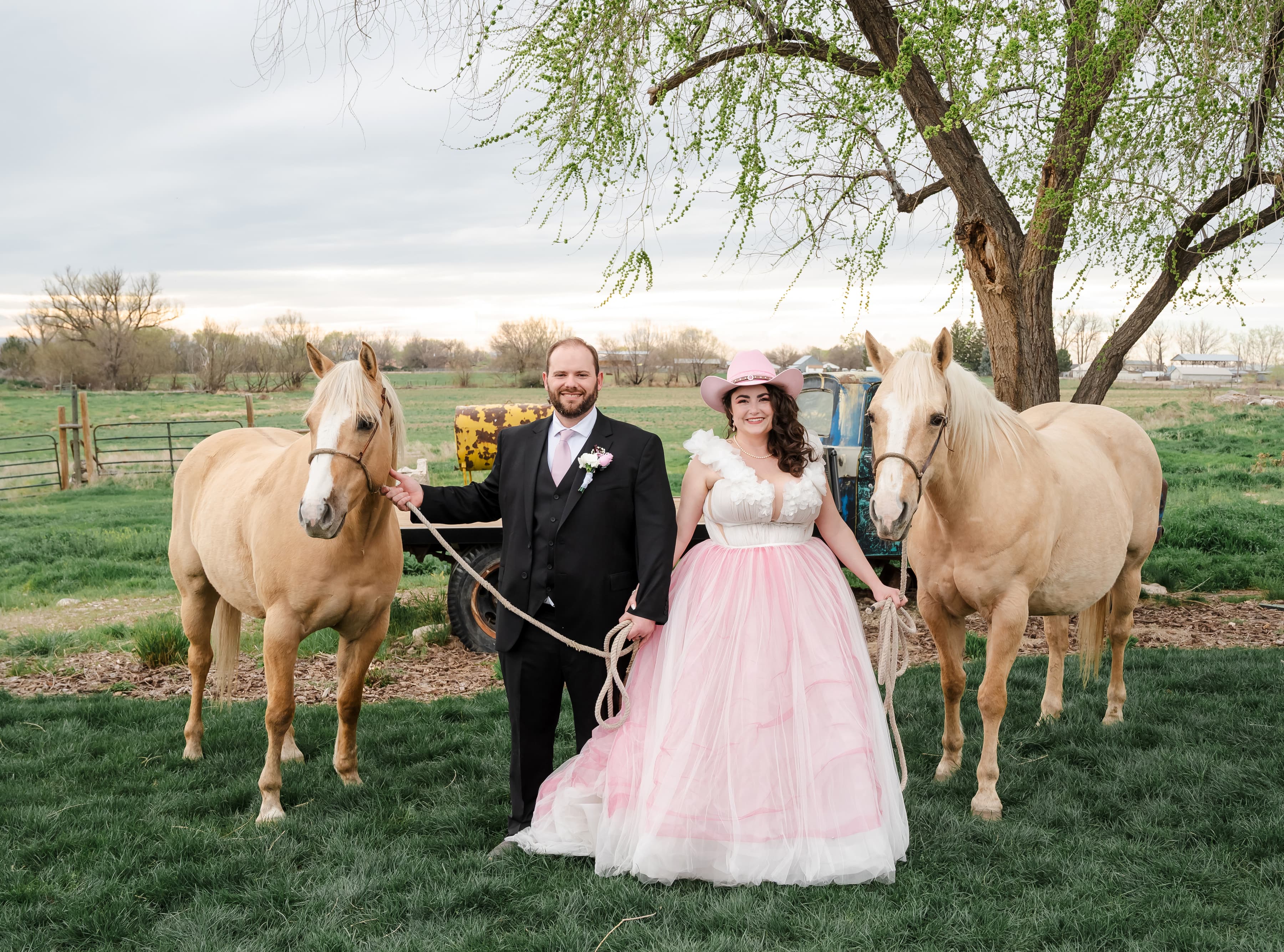 Groom and bride standing outdoors between two palomino horses. The bride is wearing a pink cowgirl hat.