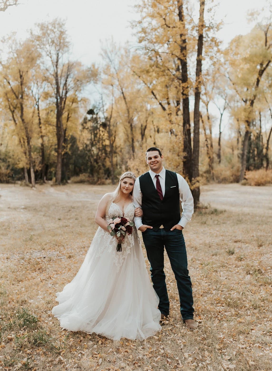 Bride and groom standing outside in the fall, behind them are trees with yellow leaves.
